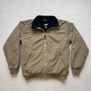 May include: A tan jacket with a black collar and a full zipper closure. The jacket has two front pockets and ribbed cuffs and hem. The brand name "Guide" is embroidered on the chest. The jacket is made of a lightweight material.