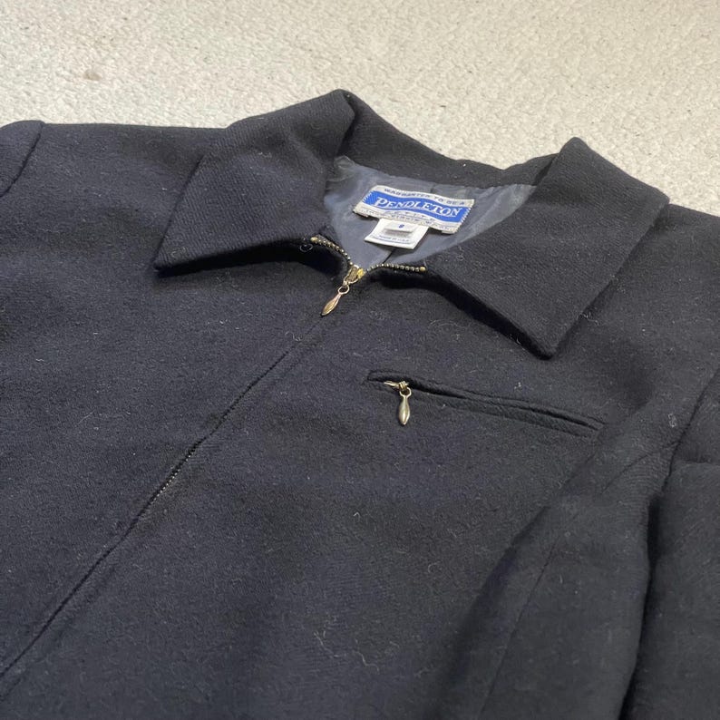 May include: A black wool jacket with a collar and a full-length zipper. The jacket has a small pocket on the chest and a label that reads "Pendleton". The jacket is a classic style.