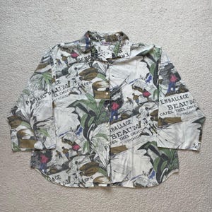 Puede incluir: Una camisa blanca de manga larga con botones con un estampado colorido que presenta plantas tropicales, granos de café y el texto "Emballage Beauté Cafés, Thés Chocolat Torrefactie".