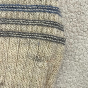 Pode incluir: Close-up de uma se&ccedil;&atilde;o de meia de malha. A meia &eacute; principalmente branco-sujo com listras horizontais azuis e cinzas. O padr&atilde;o de malha &eacute; vis&iacute;vel e a textura parece macia e quente. A meia &eacute; provavelmente feita de l&atilde; ou material semelhante.