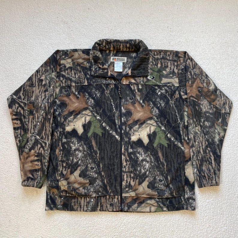 May include: A brown, green, and black camouflage fleece jacket with a zipper closure and a small logo on the chest. The jacket is made by the brand "MOOSE CREEK".