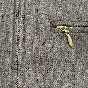 May include: Close-up of a dark gray fabric with a gold-colored zipper and a small pocket. The zipper pull is a teardrop shape. The fabric has a subtle woven texture.