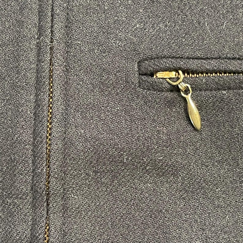 May include: Close-up of a dark gray fabric with a gold-colored zipper and a small pocket. The zipper pull is a teardrop shape. The fabric has a subtle woven texture.