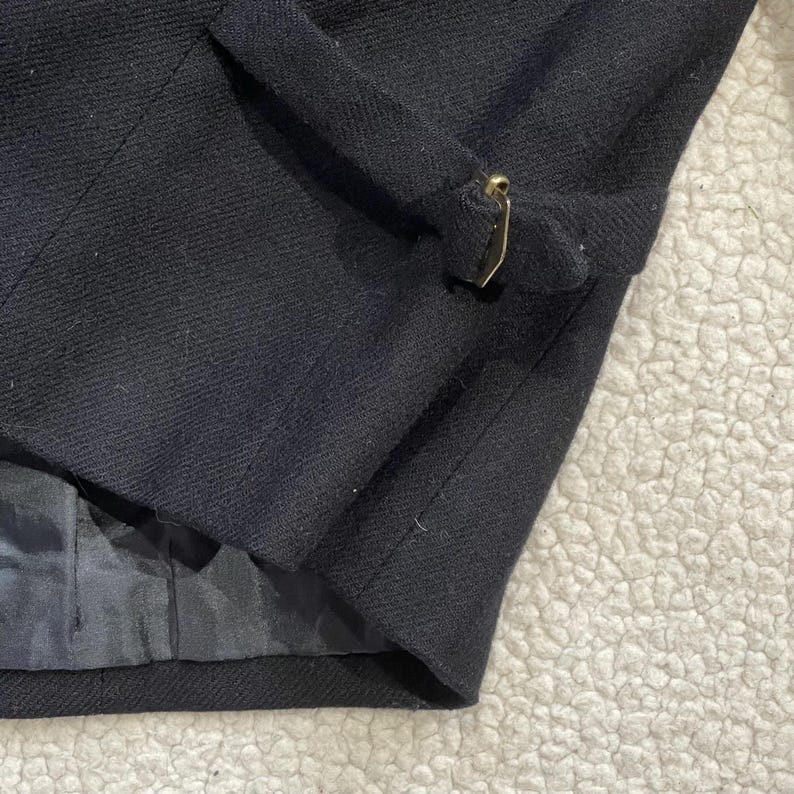 May include: Close-up of a black wool blazer with a gold-colored metal clasp. The blazer has a tailored fit and a satin lining. The fabric has a subtle texture, and the clasp is rectangular.