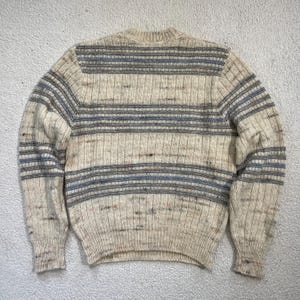 Pode incluir: Camisola creme com riscas horizontais em tons de azul e cinza. O tecido de malha apresenta uma textura canelada e gola redonda. A camisola tem mangas compridas e um estilo cl&aacute;ssico e casual.