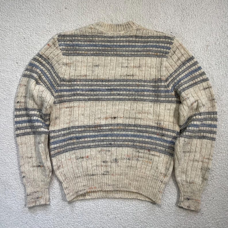 Pode incluir: Camisola creme com riscas horizontais em tons de azul e cinza. O tecido de malha apresenta uma textura canelada e gola redonda. A camisola tem mangas compridas e um estilo cl&aacute;ssico e casual.
