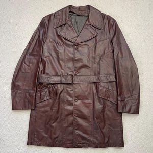 May include: A burgundy leather coat with a belted waist and a button-up front. The coat has a classic design with a notched collar and two front pockets. The coat is a medium length and appears to be vintage.