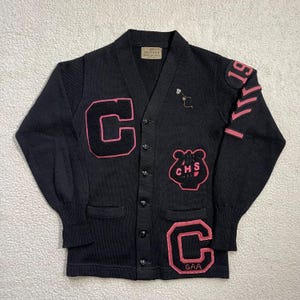Puede incluir: Cárdigan negro vintage con parches bordados en rosa. Presenta una gran "C" rosa en la parte delantera, un emblema "CHS" y un parche "GAA". La manga derecha tiene "19" y rayas. El cárdigan tiene cuello en V y cierre de botones.