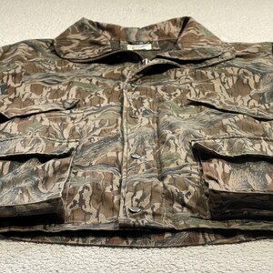 May include: Camouflage jacket with a button-up front, collar, and multiple pockets. The jacket features a pattern of brown, green, and tan hues, typical of woodland camouflage. The garment appears to be made of a durable, possibly water-resistant material.