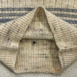 Pode incluir: Close-up de um su&eacute;ter de malha creme com textura canelada. O su&eacute;ter tem manchas de cor e listras azuis horizontais na parte superior do peito. A gola est&aacute; dobrada.