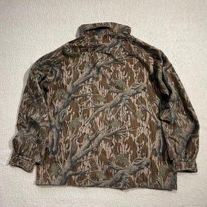 May include: A long-sleeved button-down shirt with a camouflage pattern. The shirt features a brown, tan, and green tree branch design. The collar and cuffs are visible. The shirt is made of a durable fabric.