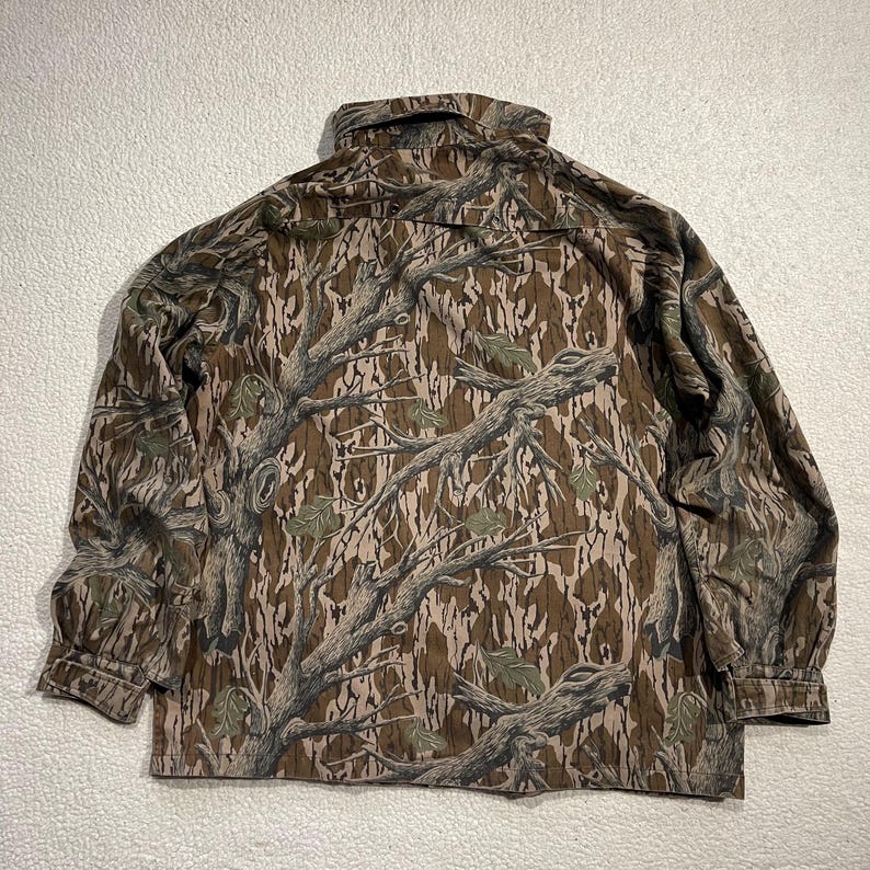 May include: A long-sleeved button-down shirt with a camouflage pattern. The shirt features a brown, tan, and green tree branch design. The collar and cuffs are visible. The shirt is made of a durable fabric.