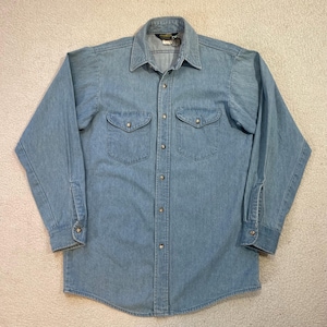 May include: Pale blue denim shirt with long sleeves, a button-down front, and two chest pockets. The shirt features a classic collar and buttoned cuffs. The fabric appears to be a medium-weight denim.