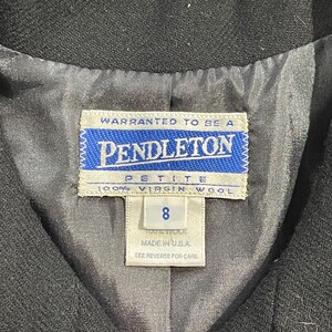 May include: Close-up of a black garment's interior, showcasing a Pendleton label. The label is white with blue text and a blue border, reading "PENDLETON PETITE 100% VIRGIN WOOL 8 MADE IN U.S.A." The lining is a light gray fabric.