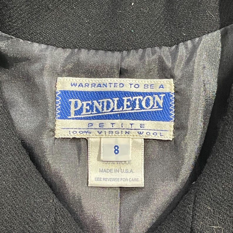 May include: Close-up of a black garment's interior, showcasing a Pendleton label. The label is white with blue text and a blue border, reading "PENDLETON PETITE 100% VIRGIN WOOL 8 MADE IN U.S.A." The lining is a light gray fabric.