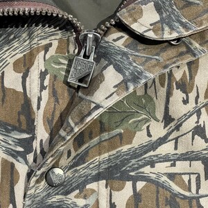 May include: Close-up of a camouflage jacket featuring a zipper with the text "North Club" and a button with a logo. The jacket has a pattern of brown, green, and gray, resembling a forest environment. The zipper is silver.