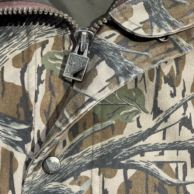 May include: Close-up of a camouflage jacket featuring a zipper with the text "North Club" and a button with a logo. The jacket has a pattern of brown, green, and gray, resembling a forest environment. The zipper is silver.