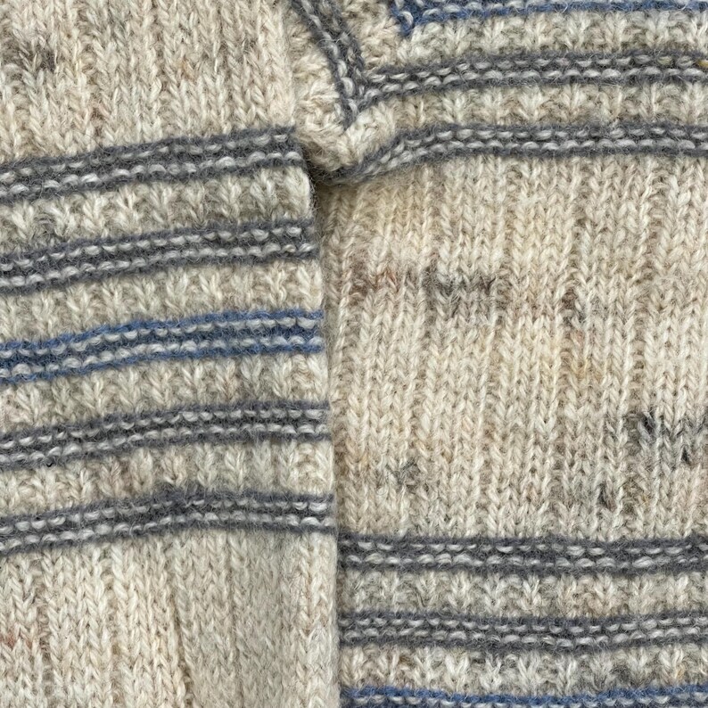 Pode incluir: Close-up de um su&eacute;ter de malha com base creme e listras horizontais cinza e azul. A malha tem uma apar&ecirc;ncia texturizada, com pontos vis&iacute;veis e uma est&eacute;tica macia e quente. O design &eacute; simples e cl&aacute;ssico.