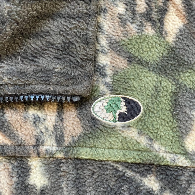 May include: A brown, green, and tan camouflage fleece jacket with a zipper closure and a small embroidered patch with a tree and a silhouette of a person's head.