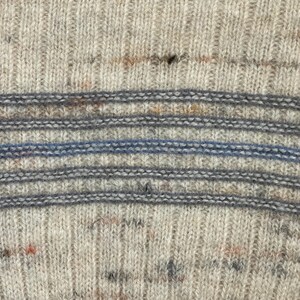 Pode incluir: Close-up de um su&eacute;ter de malha com textura canelada. O su&eacute;ter apresenta listras horizontais em tons de cinza e uma &uacute;nica listra azul. A malha tem manchas de cor, adicionando interesse visual.