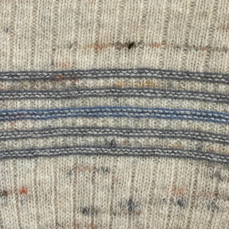 Pode incluir: Close-up de um su&eacute;ter de malha com textura canelada. O su&eacute;ter apresenta listras horizontais em tons de cinza e uma &uacute;nica listra azul. A malha tem manchas de cor, adicionando interesse visual.