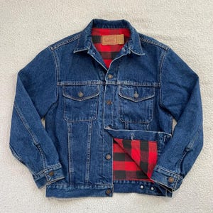 May include: A blue denim jacket with a red and black plaid flannel lining. The jacket has two chest pockets and two side pockets. The collar is a classic style, and the jacket is buttoned up. The jacket is a medium wash denim.