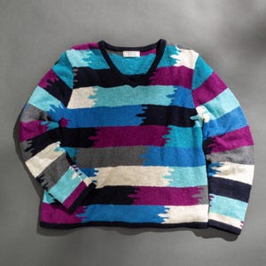 Könnte beinhalten: Ein langärmliger V-Ausschnitt-Pullover mit horizontalen Streifen in Blau-, Lila-, Grau- und Weißtönen. Der Pullover hat einen dunkelblauen Besatz. Das Muster hat einen Pinselstrich-Effekt.