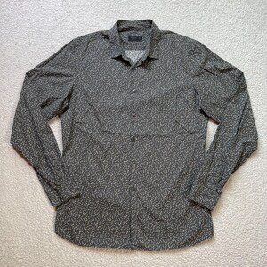 Pode incluir: Uma camisa cinza escuro com botões com um padrão branco sutil e repetitivo. A camisa de manga comprida tem uma gola clássica e punhos abotoados. O tecido parece ser um material leve.