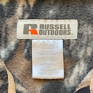 May include: A brown, black, and white camouflage jacket tag with the text "RUSSELL OUTDOORS" in white. The tag also includes information about the jacket's material, care instructions, and manufacturer.