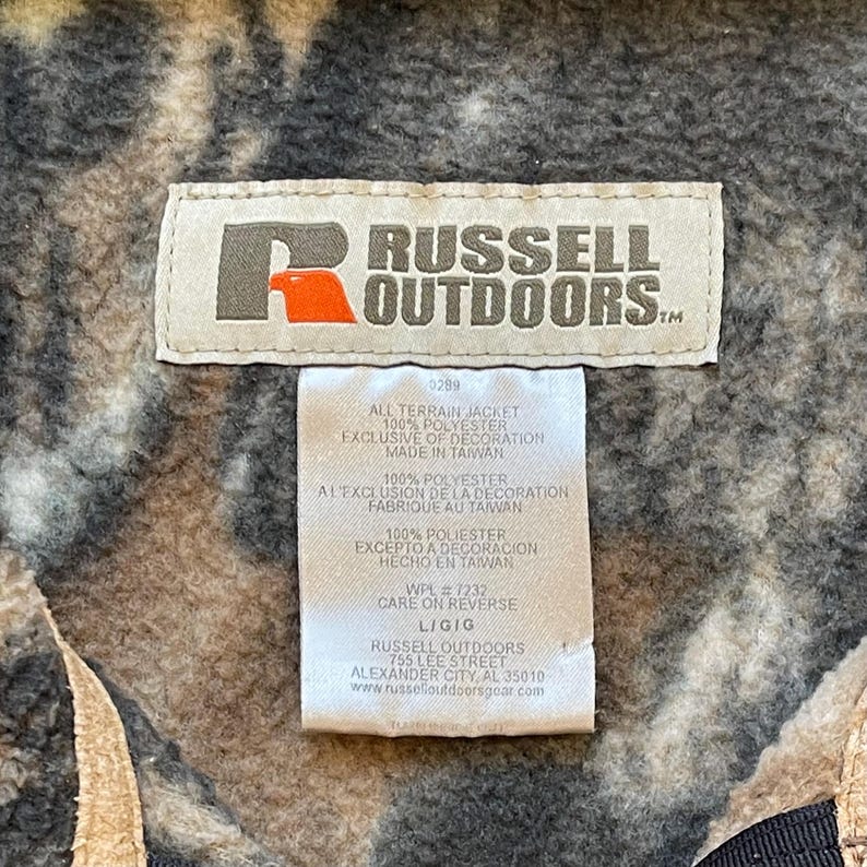 May include: A brown, black, and white camouflage jacket tag with the text "RUSSELL OUTDOORS" in white. The tag also includes information about the jacket's material, care instructions, and manufacturer.