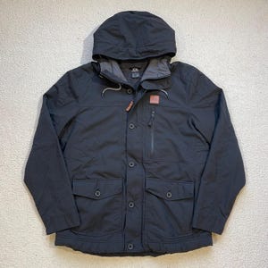 May include: A black hooded jacket with a button front closure and multiple pockets. The jacket has a logo patch on the left chest and a zippered pocket on the right side.