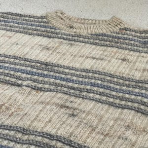 Pode incluir: Uma camisola de gola redonda com riscas horizontais em tons de bege, cinza e azul. O tecido de malha tem um aspeto texturizado, com gola canelada e um padr&atilde;o diagonal no corpo.
