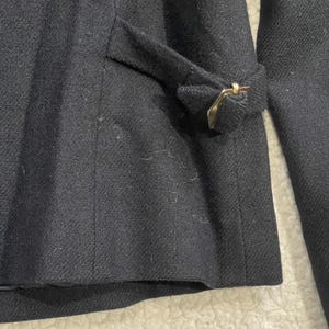 May include: Close-up of a black wool coat. The coat features a tailored design with a notched lapel and a decorative gold-colored buckle detail on the sleeve. The fabric has a subtle texture, suggesting a high-quality material.