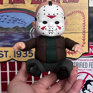 May include: A small, seated figurine of a baby wearing a Jason Voorhees mask. The figure has a brown jacket, green shirt, and gray pants. The mask is white with black eye holes and red accents. The figurine is held in a hand.