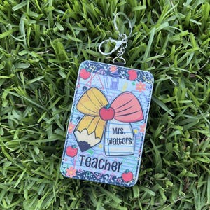 May include: A rectangular teacher keyring featuring a pencil, apple, and bow design. The keyring has the text "Teacher" and "Mrs. Walters". The background is light blue with school-themed graphics. It is attached to a silver keyring.