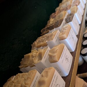May include: A display of handcrafted soaps in various shades of beige and brown, each in a white paper sleeve. The soaps are arranged on a wooden shelf, with some labeled "Eucalyptus & Spearmint". The soaps have a textured surface.