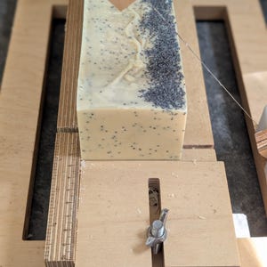 May include: A rectangular block of handmade soap with poppy seeds is being cut with a wooden soap cutter. The soap is light beige with dark gray poppy seeds. The cutter is made of light-colored wood and has a metal wire for slicing.
