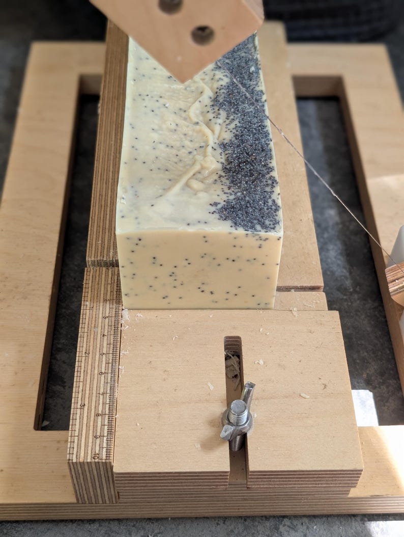 May include: A rectangular block of handmade soap with poppy seeds is being cut with a wooden soap cutter. The soap is light beige with dark gray poppy seeds. The cutter is made of light-colored wood and has a metal wire for slicing.