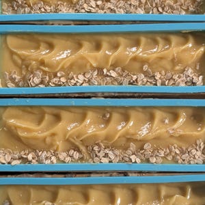 May include: Close-up of several bars of handmade soap in a blue mold. The soap is a light tan color with a textured surface and embedded oats along the bottom edge. The soap appears to be freshly made.