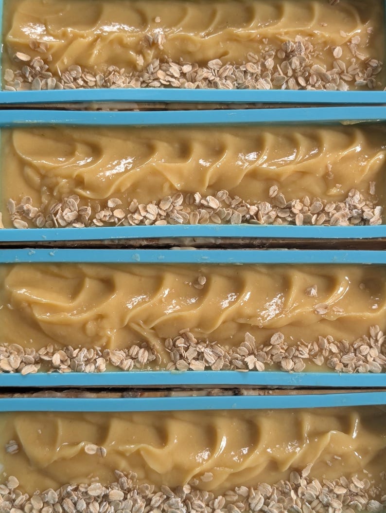 May include: Close-up of several bars of handmade soap in a blue mold. The soap is a light tan color with a textured surface and embedded oats along the bottom edge. The soap appears to be freshly made.