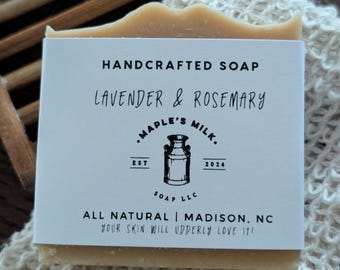 May include: A bar of handcrafted soap with a white label that reads "Lavender & Rosemary." The label also includes the text "Maple's Milk Soap LLC" and "All Natural | Madison, NC." The soap is light brown and rests on a beige washcloth.
