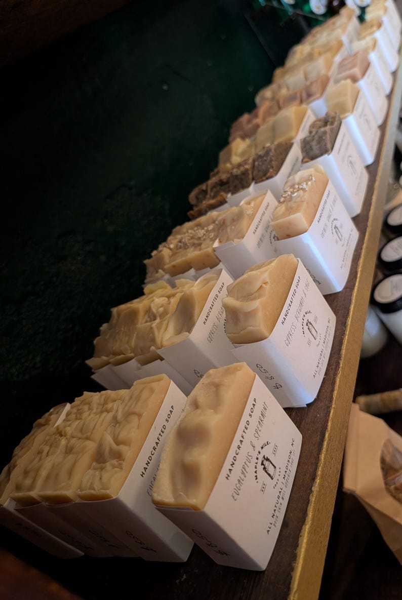 May include: A display of handcrafted soaps in various shades of beige and brown, each in a white paper sleeve. The soaps are arranged on a wooden shelf, with some labeled "Eucalyptus & Spearmint". The soaps have a textured surface.