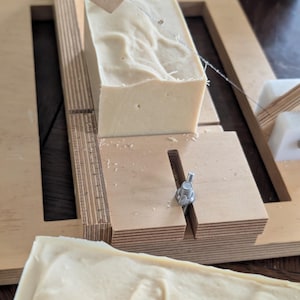May include: A light beige rectangular bar of soap being cut with a wooden soap cutter. The cutter has a wire blade and a wooden frame. The soap has a textured surface and is ready to be sliced into smaller bars. The wooden cutter has a ruler.