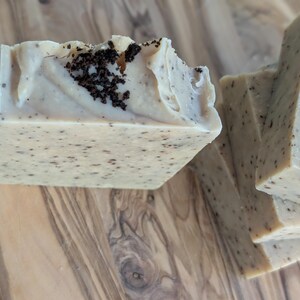 May include: Close-up of several handmade soap bars. The soap is a light beige color with dark speckles and a textured top. Some bars have a dark, granular topping. The soaps are arranged on a wooden surface.
