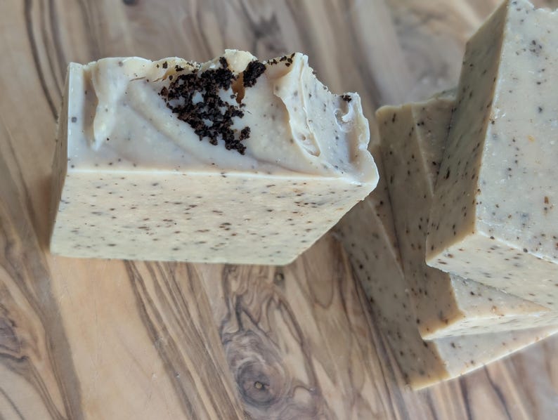 May include: Close-up of several handmade soap bars. The soap is a light beige color with dark speckles and a textured top. Some bars have a dark, granular topping. The soaps are arranged on a wooden surface.