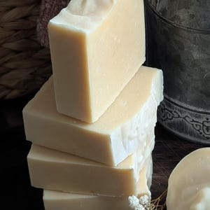 May include: A stack of four rectangular bars of handmade soap, with a light beige color and a slightly textured surface. A small sprig of white flowers rests near the soap. A round soap and a metal bucket are also in the image.