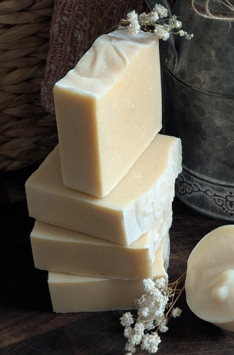 May include: A stack of four rectangular bars of handmade soap, with a light beige color and a slightly textured surface. A small sprig of white flowers rests near the soap. A round soap and a metal bucket are also in the image.