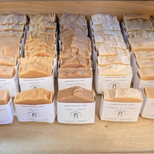 May include: A wooden tray filled with various handcrafted soaps in white paper packaging. The soaps are in shades of beige, tan, and brown. Each package has a label with text and a logo. The soaps are arranged in neat rows.