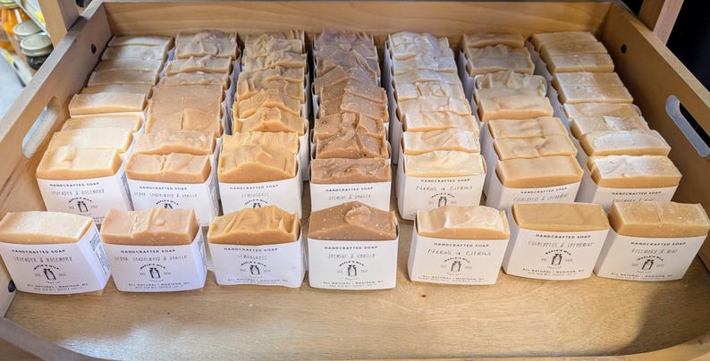 May include: A wooden tray filled with various handcrafted soaps in white paper packaging. The soaps are in shades of beige, tan, and brown. Each package has a label with text and a logo. The soaps are arranged in neat rows.
