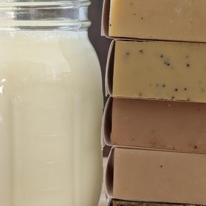 May include: A mason jar filled with a creamy liquid sits next to a stack of wrapped soap bars. The soaps vary in color, from light beige to dark brown, with some containing visible seeds or particles. The jar has measurement markings.